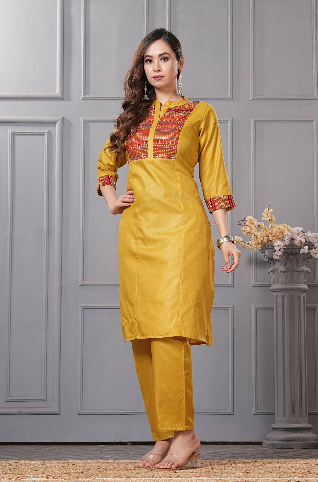 A full-length standing pose of a woman in a mustard-yellow ethnic ensemble - paired with transparent block heels - against a grey paneled background.