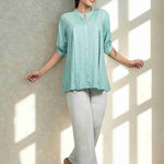 Front view of women's sea green rayon shirt featuring a mandarin collar, button-down front, front pleats, half sleeves, and a relaxed fit paired with white trousers.