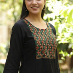 A portrait of a woman wearing a black ethnic kurta with mirror-work, set against a blurred green background.
