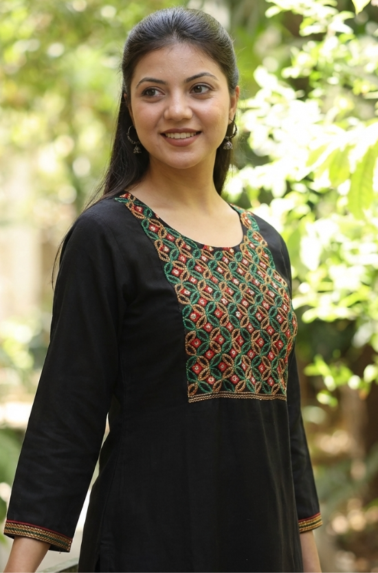 A portrait of a woman wearing a black ethnic kurta with mirror-work, set against a blurred green background.