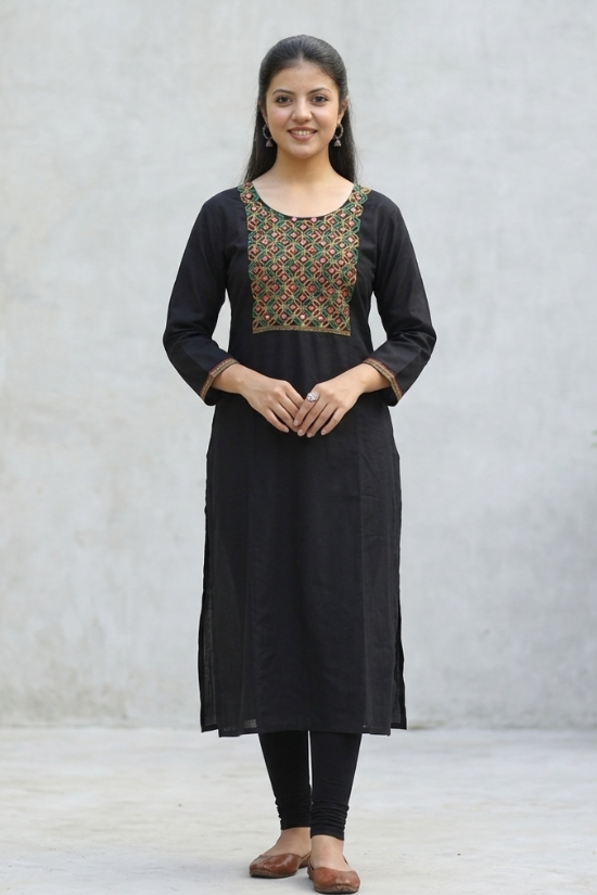 A woman leaning against a stone wall, wearing a black straight kurta with mirror-work embroidery.
