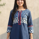 Smiling woman wearing a dark blue kurti featuring a geometric and floral printed yoke with red piping.