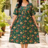 Full-length front view of a woman wearing a dark green pure cotton frock-style kurti featuring an all-over vibrant red and cream floral print, styled in a relaxed, flared silhouette.