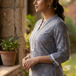 A side-profile portrait of a woman standing near a stone wall, modeling a grey-blue printed ethnic kurta.