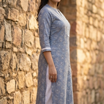 A side-profile portrait of a woman standing near a stone wall, modeling a grey-blue printed ethnic kurta.
