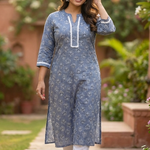 A woman walking gracefully in a garden wearing a grey-blue floral printed kurta and white straight pants.