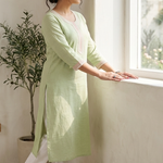 A woman standing sideways near a window, showcasing a light green kurta with intricate pink thread work and white pants.