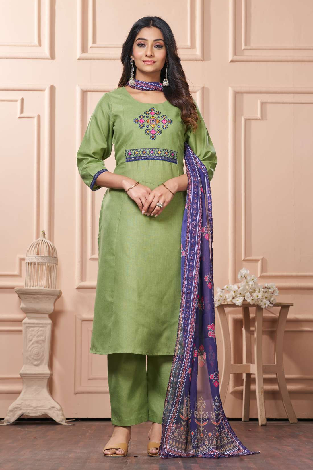 A full length presentation of a green cotton ethnic suit set displaying vibrant cross stitch embroidery details along the front panel, styled with straight pants and a printed dupatta