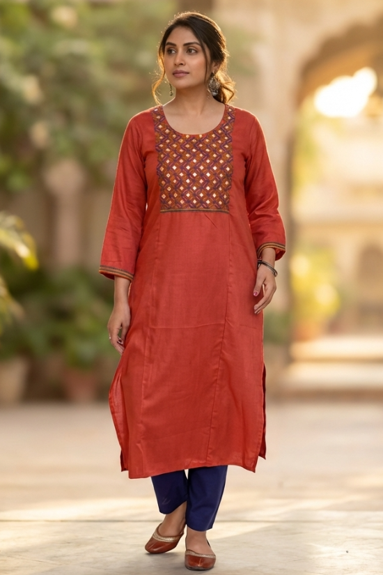 A woman walking gracefully in a garden wearing a rust-orange ethnic kurta and blue trousers.