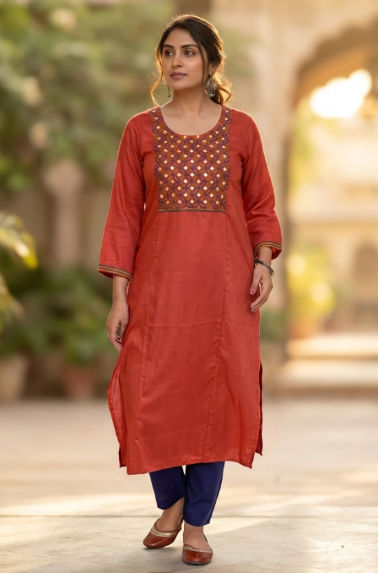A woman walking gracefully in a garden wearing a rust-orange ethnic kurta and blue trousers.