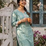A woman holding a clay cup while leaning against a wooden gate, dressed in a sea-green ethnic kurta.