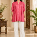 Full-length front view of a woman wearing a dark peach rayon pleated tunic top paired with white straight-leg trousers.