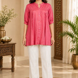 Full-length front view of a woman wearing a dark peach rayon pleated tunic top paired with white straight-leg trousers.